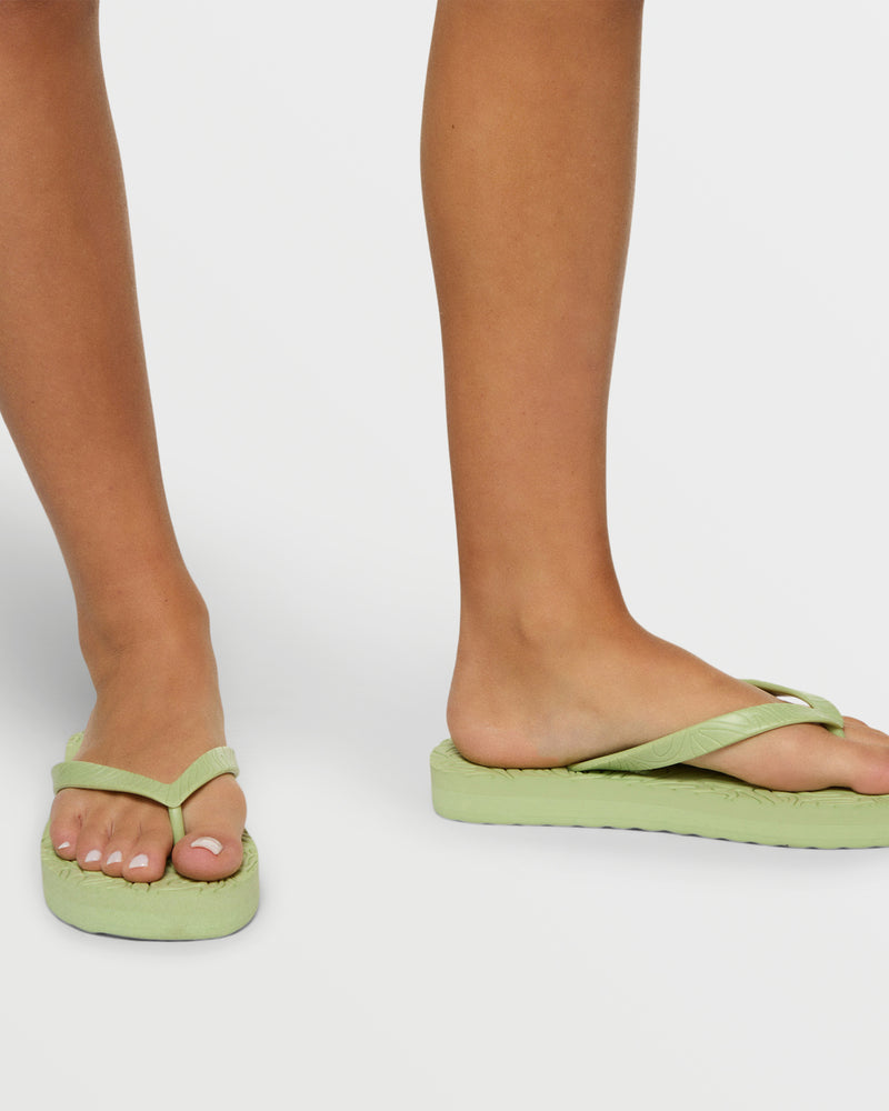 A close-up of a person with light skin wearing Roxy Beach Up Sandal in light green, showing their lower legs and feet with neatly painted white toenails against a plain white background, highlighting the stylish beach footwear.
