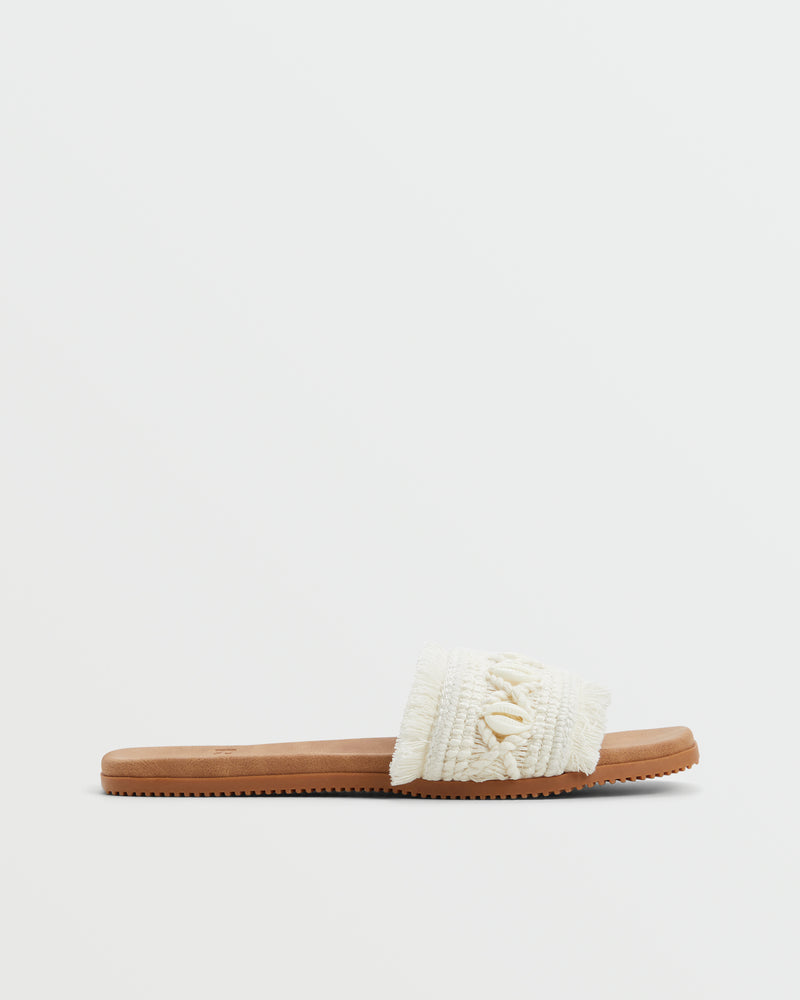 The Roxy Positano Sandal is a flat sandal with a tan sole and a wide, textured white fabric strap with fringe and decorative accents, shown against a plain white background.