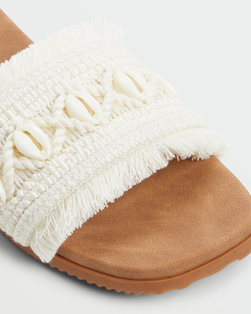 Close-up of the Roxy Positano Sandal featuring a tan sole and a wide white fabric strap with braided accents, fringe, and small white cowrie shells.