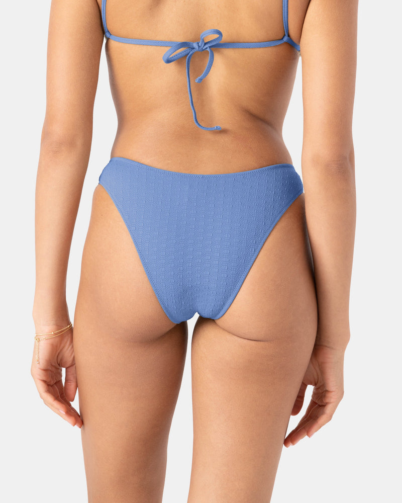 A woman is seen from the back wearing a Roxy Haleiwa Scoop Cheeky Bikini Bottom and a matching tie-back bikini top, both in textured swim fabric. She accessorizes with a gold bracelet on her left wrist against a plain white background.