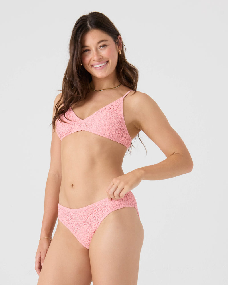 A woman with long brown hair smiles while wearing the Roxy Crochet Hipster Full Bikini Bottom in light pink, standing against a plain light gray background with one hand resting on her hip.