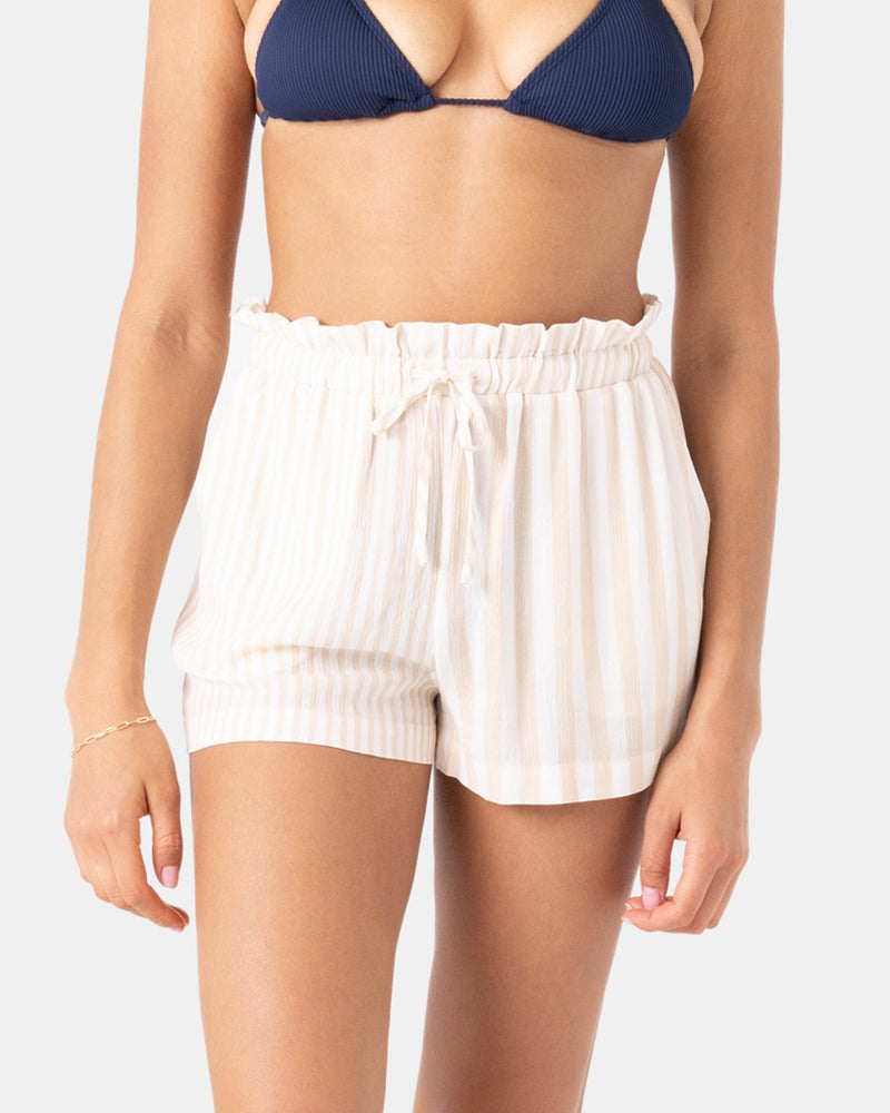 A woman stands against a light background wearing Roxy's Stripe Out Cover Up Short, featuring a textured print, vertical beige stripes, and a drawstring waist, paired with a navy blue bikini top.