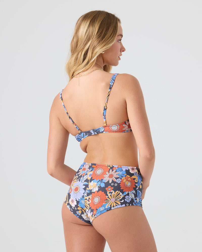 A woman with long, wavy blonde hair is seen from behind wearing the Roxy Soul Flower Boyleg Bikini Bottom, featuring a matching floral print in orange, blue, and white flowers on a light gray background.