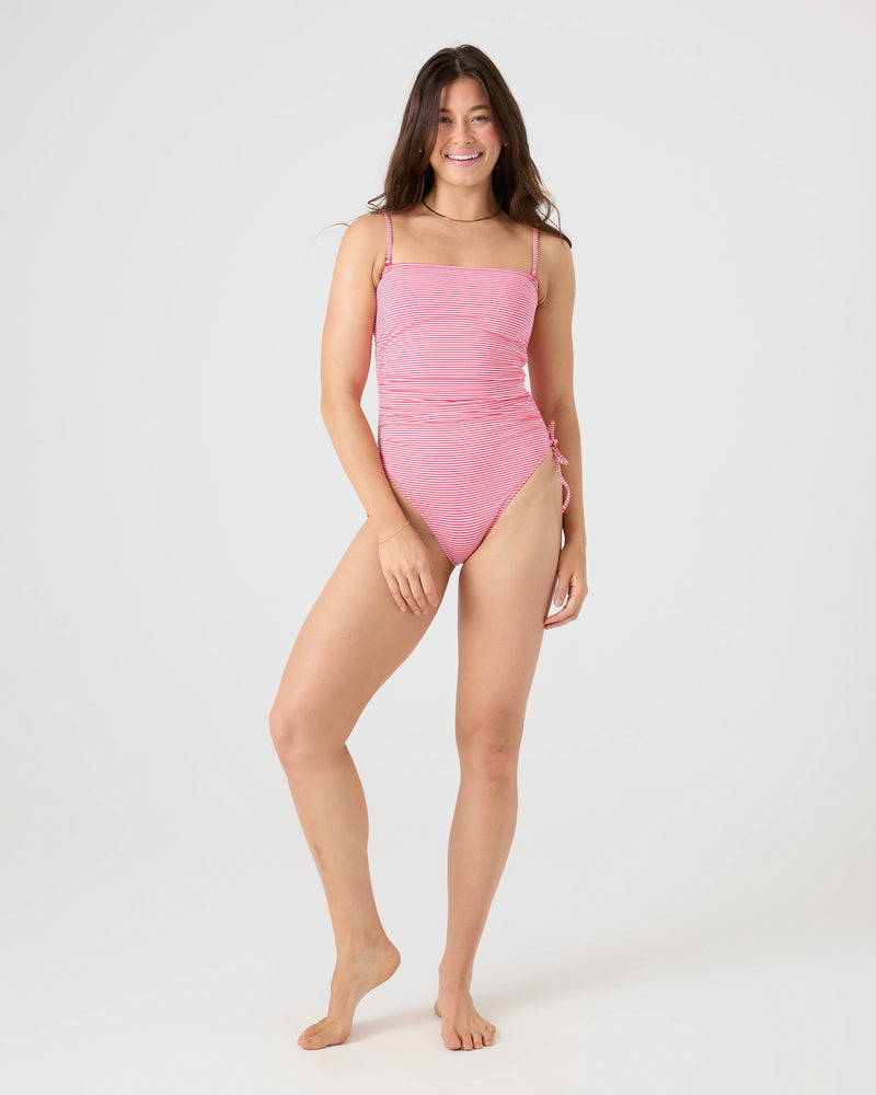 A woman with long brown hair smiles while modeling the Roxy Good Vibes Adjustable One Piece Moderate Swimsuit in pink, standing barefoot against a plain white background with one hand by her side and the other slightly bent.