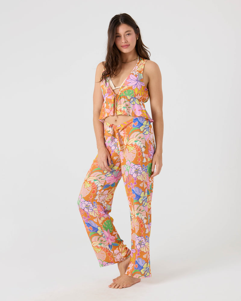 A woman stands barefoot in the Roxy Lei It On Me Cover Up Pant, featuring a colorful Brown Multi floral print and paired with a sleeveless ruffled top—perfect for beachwear or as a stylish cover-up, set against a plain white background.