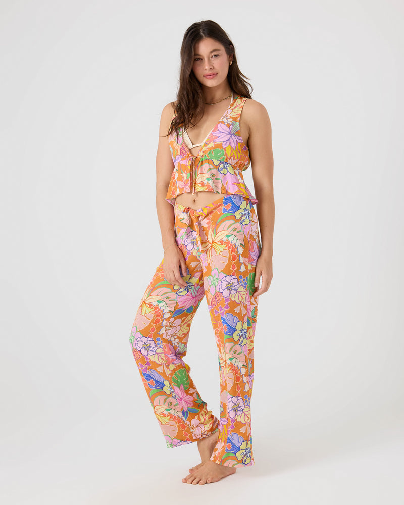 A woman stands barefoot in the Roxy Lei It On Me Cover Up Top, a colorful floral cropped tie-front sleeveless ruffled piece, with matching pants, against a plain white background.