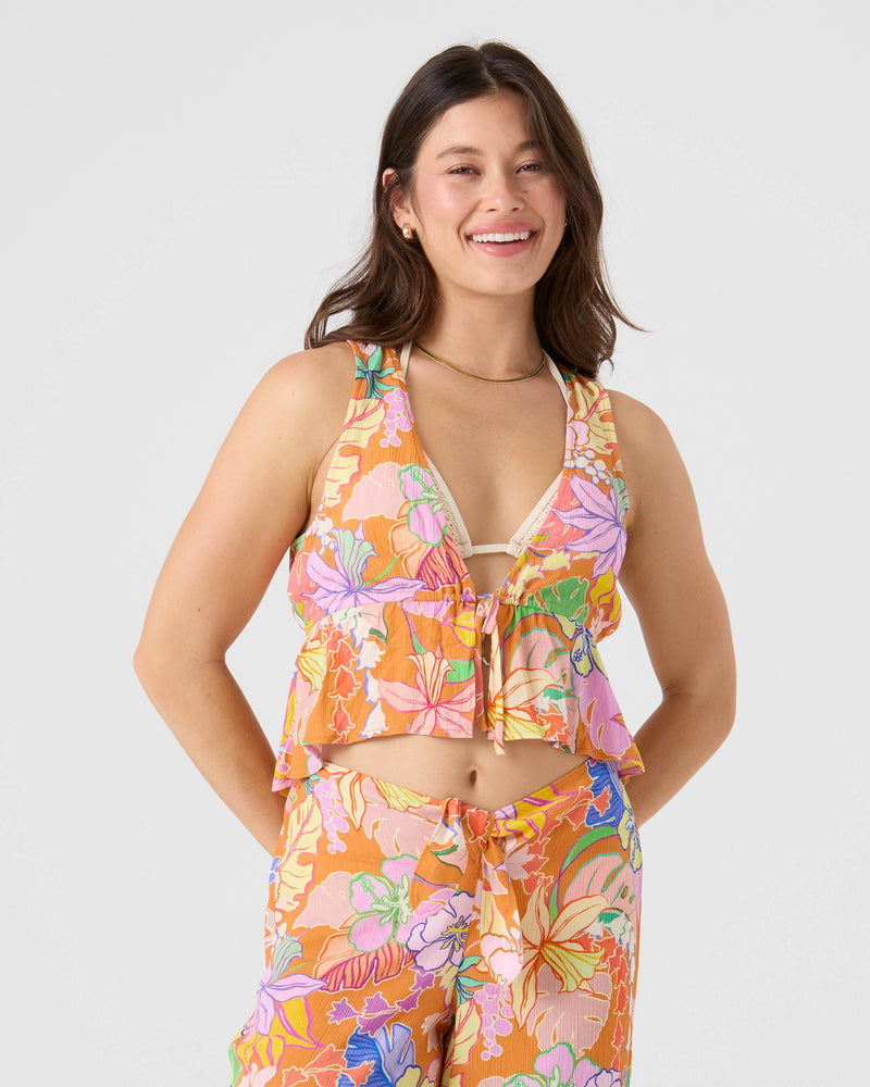 A woman with long brown hair smiles while wearing the Roxy "Lei It On Me" Cover Up Top, a colorful, floral-patterned beachwear piece in pink, orange, blue, and green hues. She stands against a plain light background.