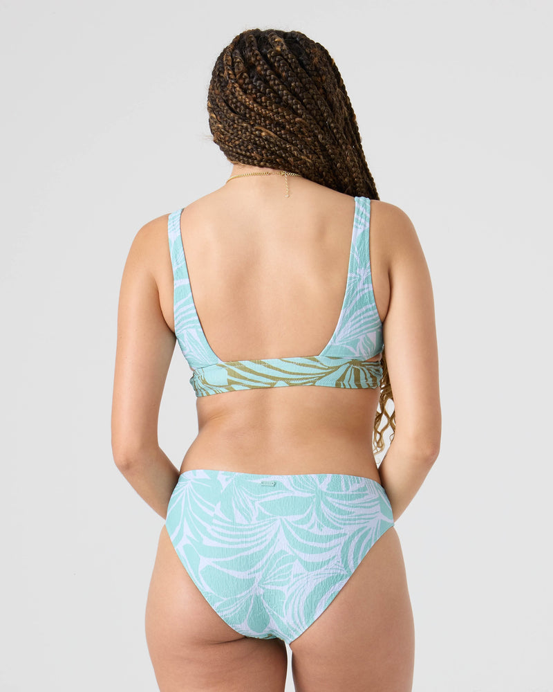 A woman with long braided hair is seen from the back wearing the Roxy Sea Bloom Cut Out Bralette Bikini Top in light blue and green patterns, featuring moderate support, set against a plain light gray background.