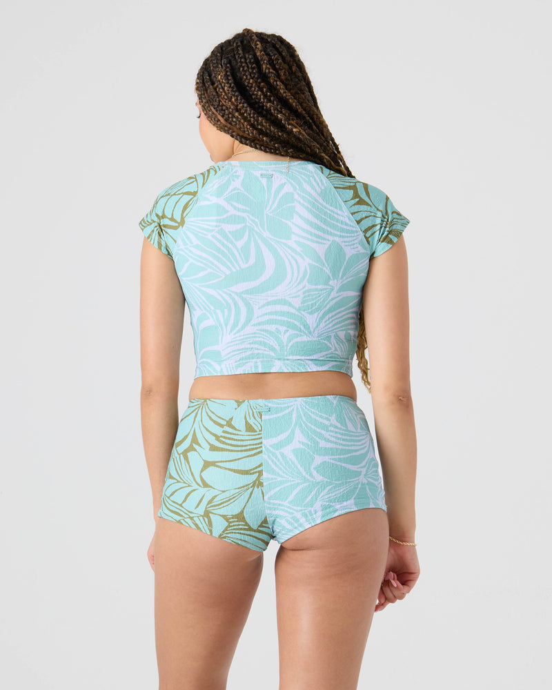 A person with long braids wears the Roxy Sea Bloom Crop Rashguard and matching shorts in a light blue and green tropical print, standing with their back to the camera against a plain background.