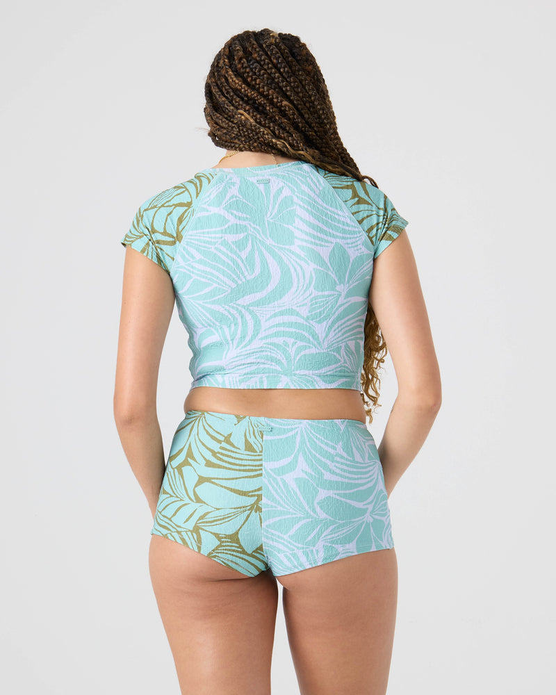Seen from behind, a person with long braided hair wears the Roxy Sea Bloom Crop Rashguard set—a short-sleeved Lycra surfwear crop and shorts in a blue-green tropical leaf pattern—standing out against a plain light gray background.