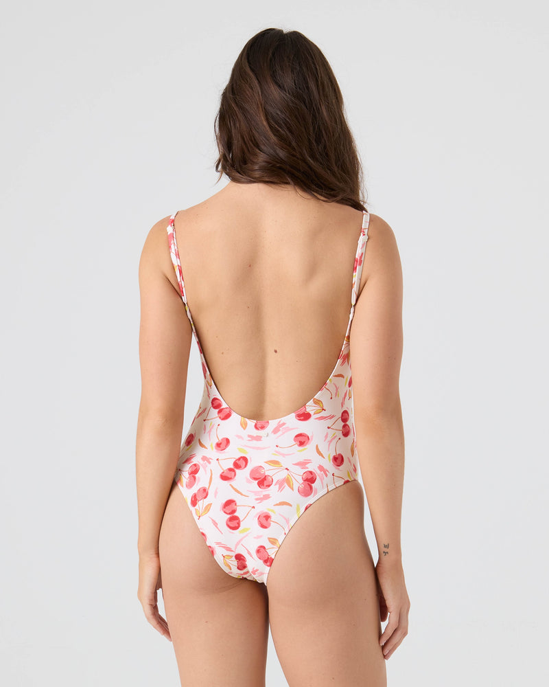 A woman with long brown hair stands with her back to the camera, wearing the Roxy Cherry On Top Low Back One Piece Swimsuit—a high-leg, white suit with a pink and orange fruit pattern, made from recycled nylon. The background is light gray.