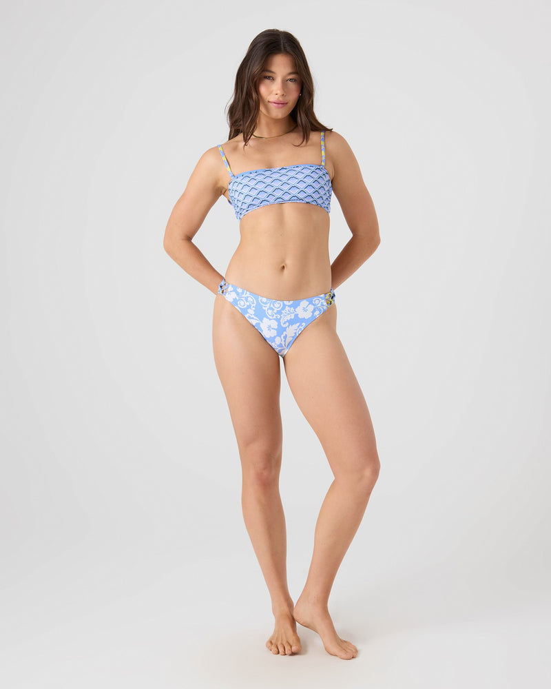 A woman with long brown hair faces forward against a plain background, wearing the Roxy Golden Glow Lace Up Bandeau Bikini Top in light blue with high-cut bottoms and a crop-top style, made from recycled nylon.