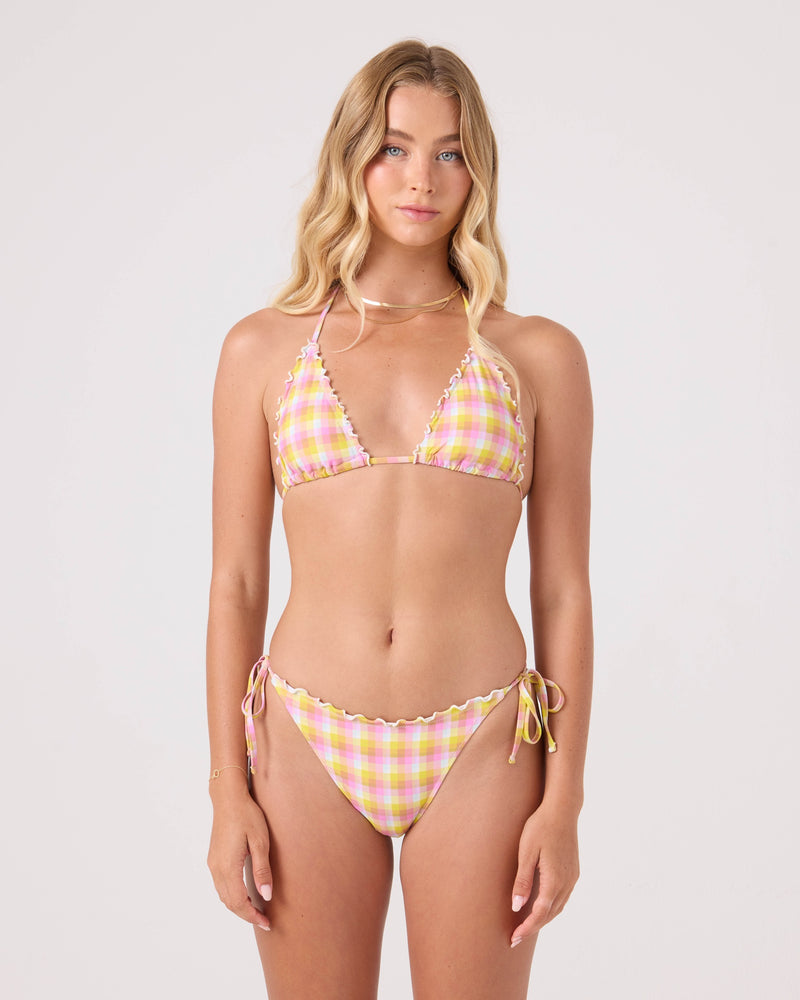 A woman with long blonde hair models the Roxy Check Me Out Mini Tiki Triangle With Ruffle Edge Bikini Top, featuring yellow-pink plaid and ruffled edges, made from recycled nylon, against a plain light background.