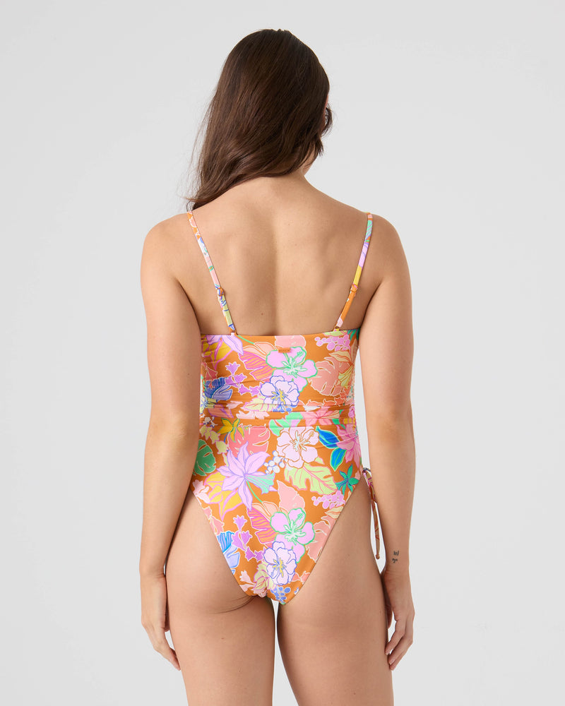 A woman with long brown hair, seen from behind, wears the Roxy Lei It On Me Adjustable Side One Piece Swimsuit—a vibrant floral design in pink, orange, and green—against a plain, light background.
