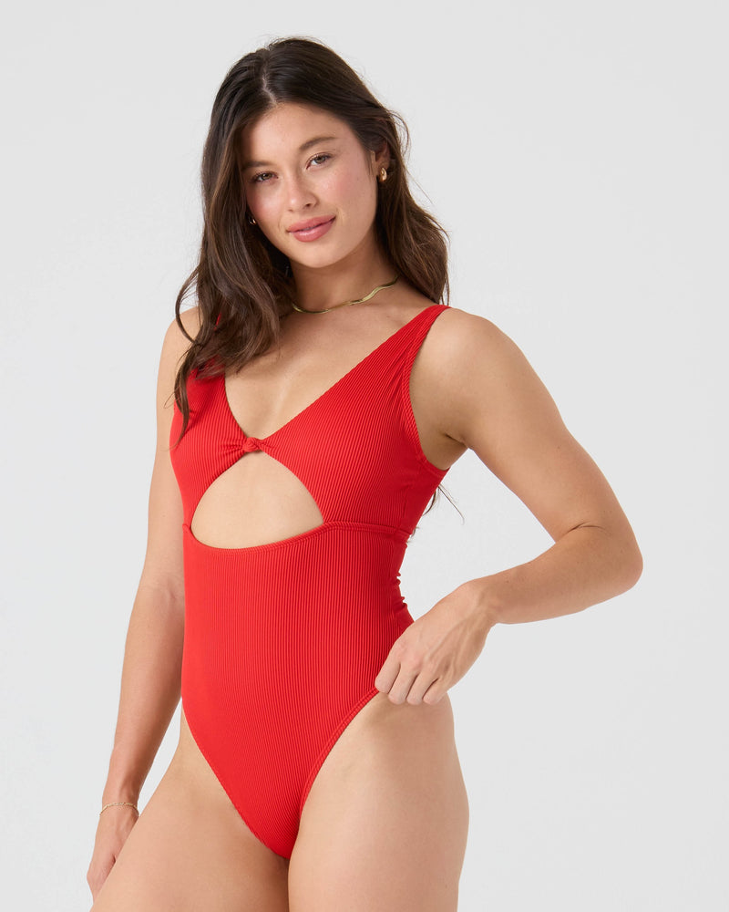 A woman with long brown hair models the Roxy Rib Love Knot Peek A Boo One Piece Moderate Swimsuit in bright red, featuring a front cutout, standing against a light gray background and smiling at the camera.