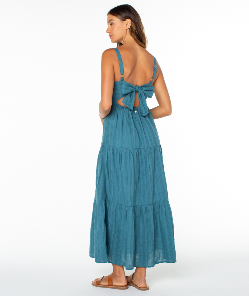 A woman faces away, wearing the Roxy Clearwater Maxi Dress in teal with a large back bow and cutout. She has long brown hair and tan flat sandals, posing against a white background.