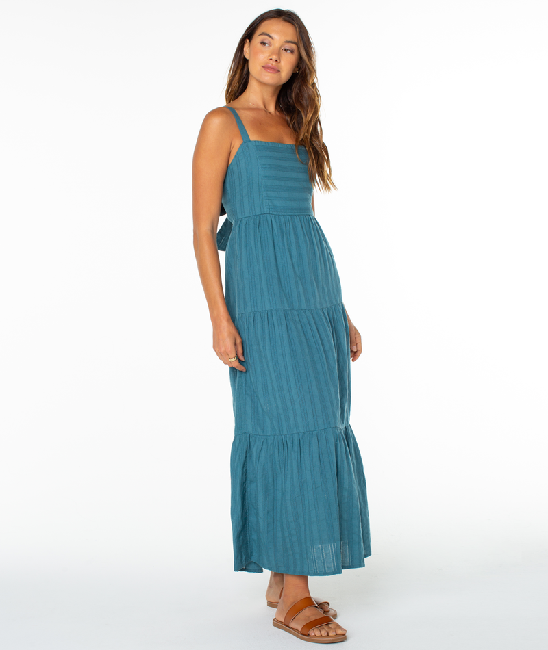 A woman with long wavy hair and a neutral expression stands against a white background in Roxy's Clearwater Maxi Dress, a sleeveless teal tiered style with layered details, paired with tan sandals.