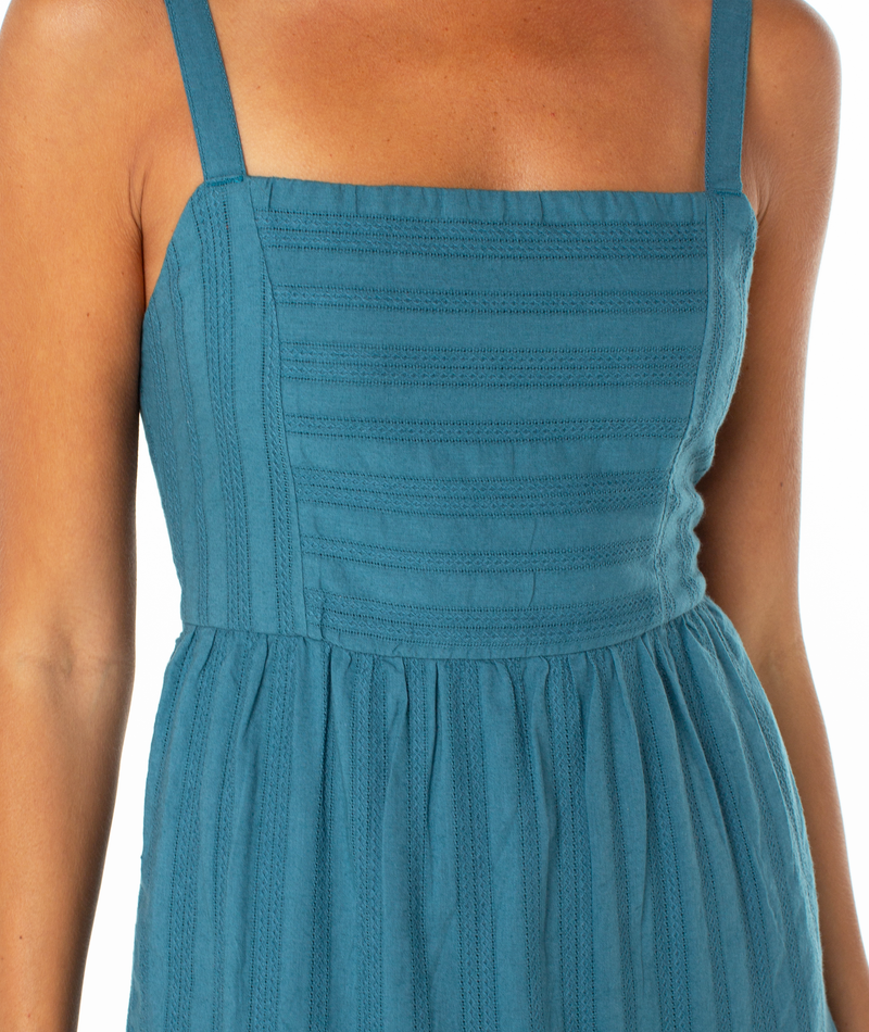 A close-up of the Roxy Clearwater Maxi Dress, a sleeveless teal piece featuring thin straps, a square neckline, textured vertical stripes, and a gathered waist. The person's arms and upper chest are partially shown.