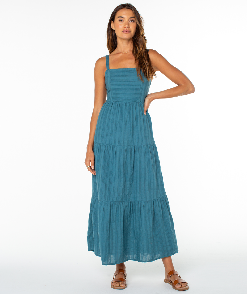 A woman with long brown hair poses against a white background in the sleeveless teal Clearwater Maxi Dress by Roxy, paired with sandals. One hand is on her hip and she has a relaxed expression.