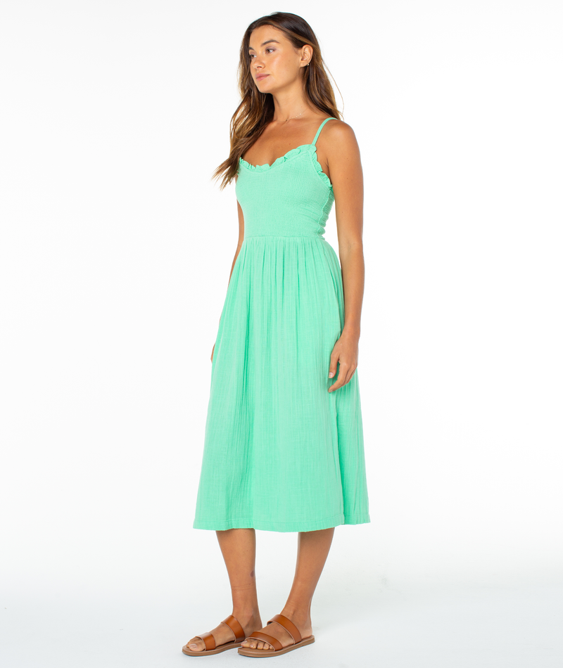 A woman stands against a white background wearing the Roxy Downriver Midi Dress—a light green, coastal style sleeveless V-neck dress with thin straps—paired with tan sandals. She faces slightly to the side, arms relaxed.