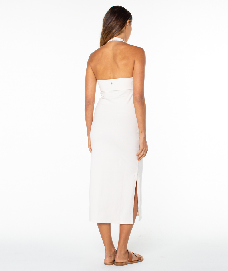 A woman with long brown hair faces away from the camera, wearing the Roxy South Shore Midi Dress—an ankle-length, strapless white dress with a side slit—paired with brown sandals against a white background.