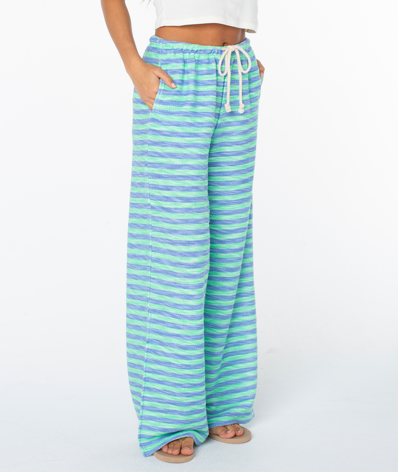 A person wears Roxy Beach Breeze Pant—loose-fitting, striped in blue, green, and purple with a white drawstring—paired with a cropped white top and sandals. Hands are in pockets, set against a plain white background.