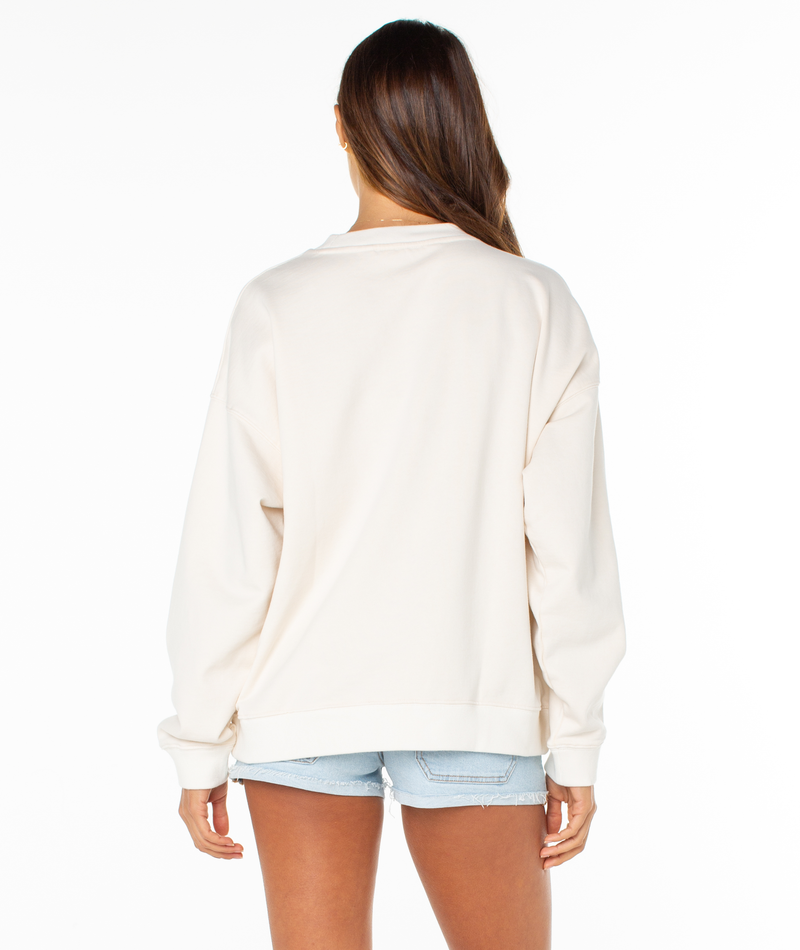 A woman with long brown hair faces away, modeling the Roxy Lineup Oversized Crew with a sun-washed finish and light blue denim shorts, set against a plain white background.