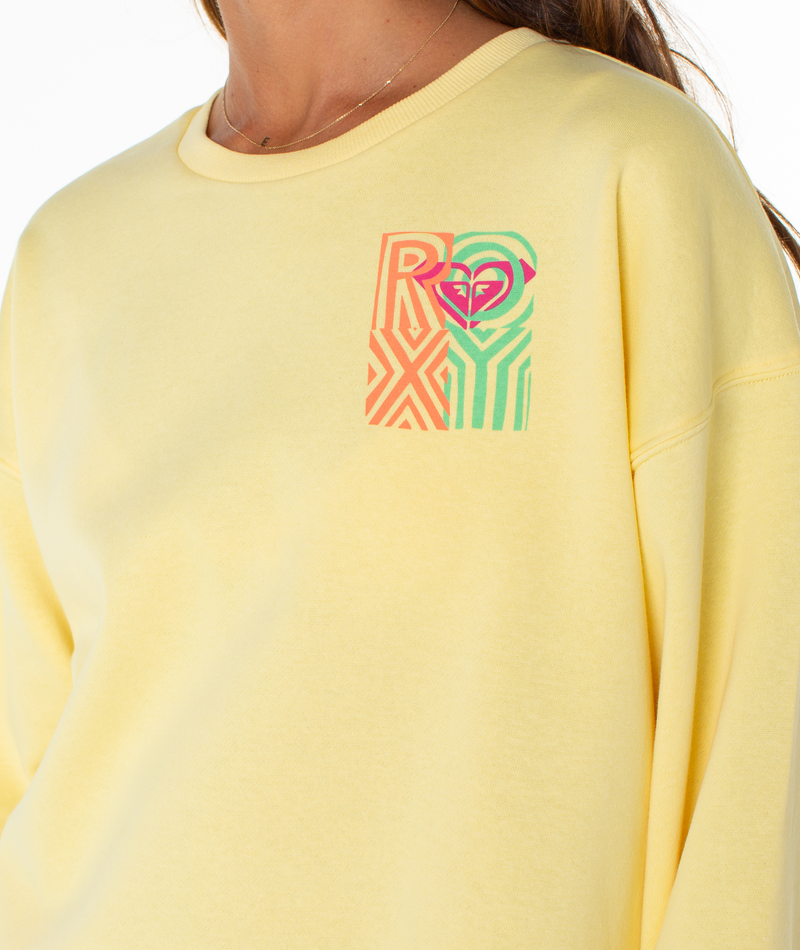 A person wearing the Roxy Morning Stroll Top, a light yellow crewneck sweatshirt featuring a colorful ROXY logo and heart graphic on the upper left chest. Only their lower face, neck, and upper body are visible.