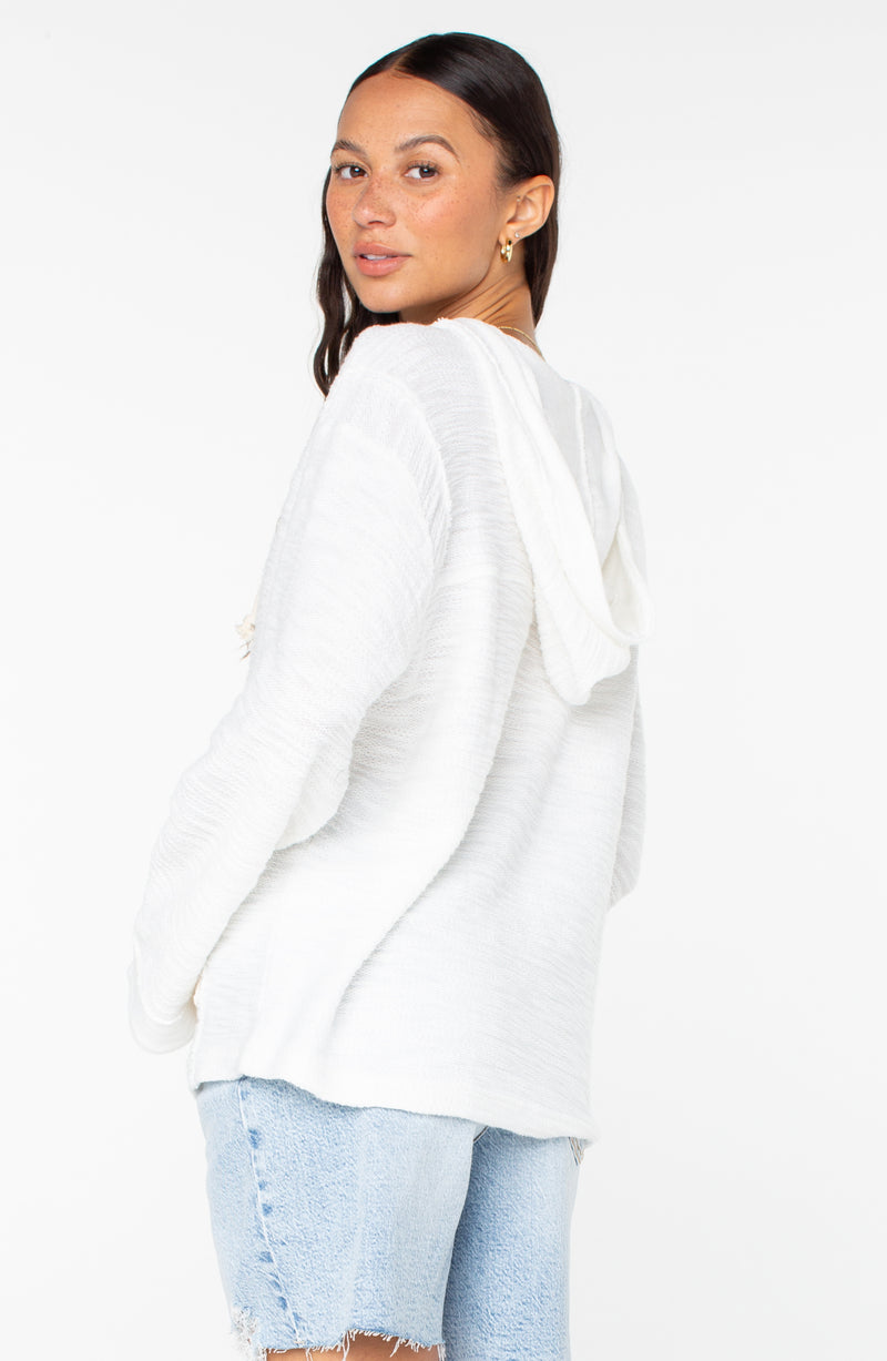A woman with long dark hair wears a white Roxy Pearling Poncho 3 made from lightweight fabric and light blue denim shorts, standing sideways and looking over her shoulder against a plain white background.