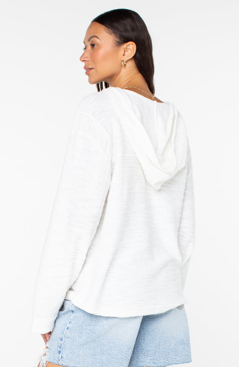 A woman with long dark hair, gold hoop earrings, and light blue denim shorts stands slightly turned against a white background, wearing the Roxy Pearling Poncho 3—an ideal lightweight French terry layer for cool evenings.