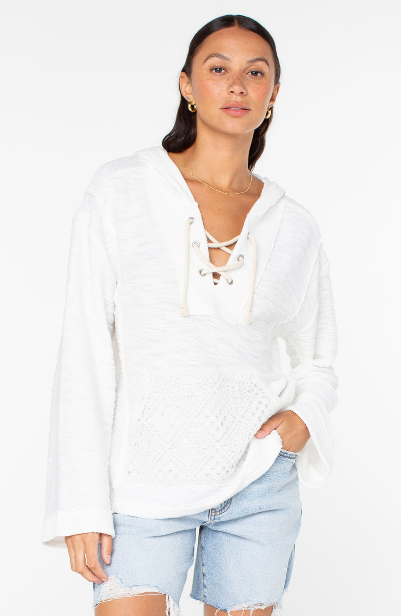 A woman wears the Roxy Pearling Poncho 3—a white, long-sleeve lace-up French terry hoodie with front pocket—paired with light blue distressed denim shorts, standing against a plain white background.