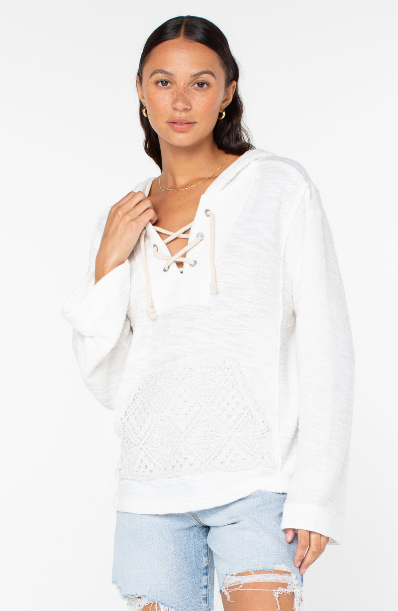 A woman wears the Roxy Pearling Poncho 3 white lace-up hoodie with front pocket and light blue distressed denim shorts, standing against a plain white background and looking at the camera.