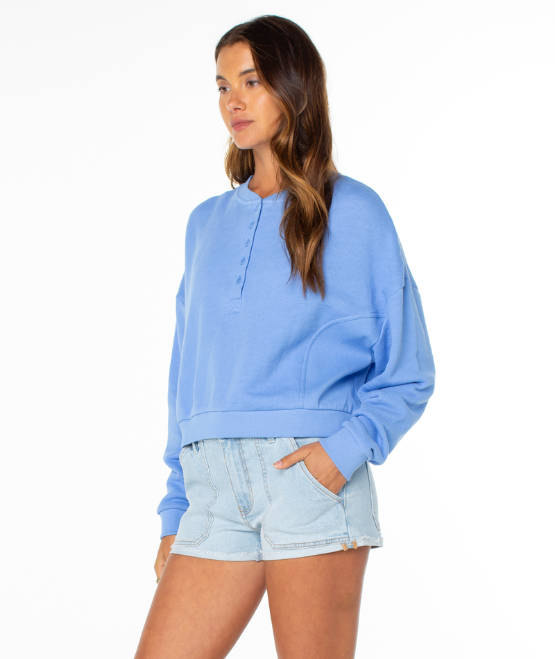 A woman stands against a white background wearing the Roxy Overcast Hoodie—a light blue long-sleeve piece—paired with light denim shorts, her hands in her pockets and hair down.