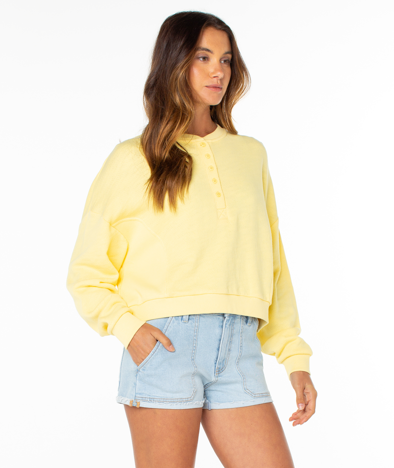 A woman stands against a white background wearing the loose-fitting yellow Roxy Overcast Hoodie with a button front and light blue denim shorts, her left hand in her pocket and hair down.