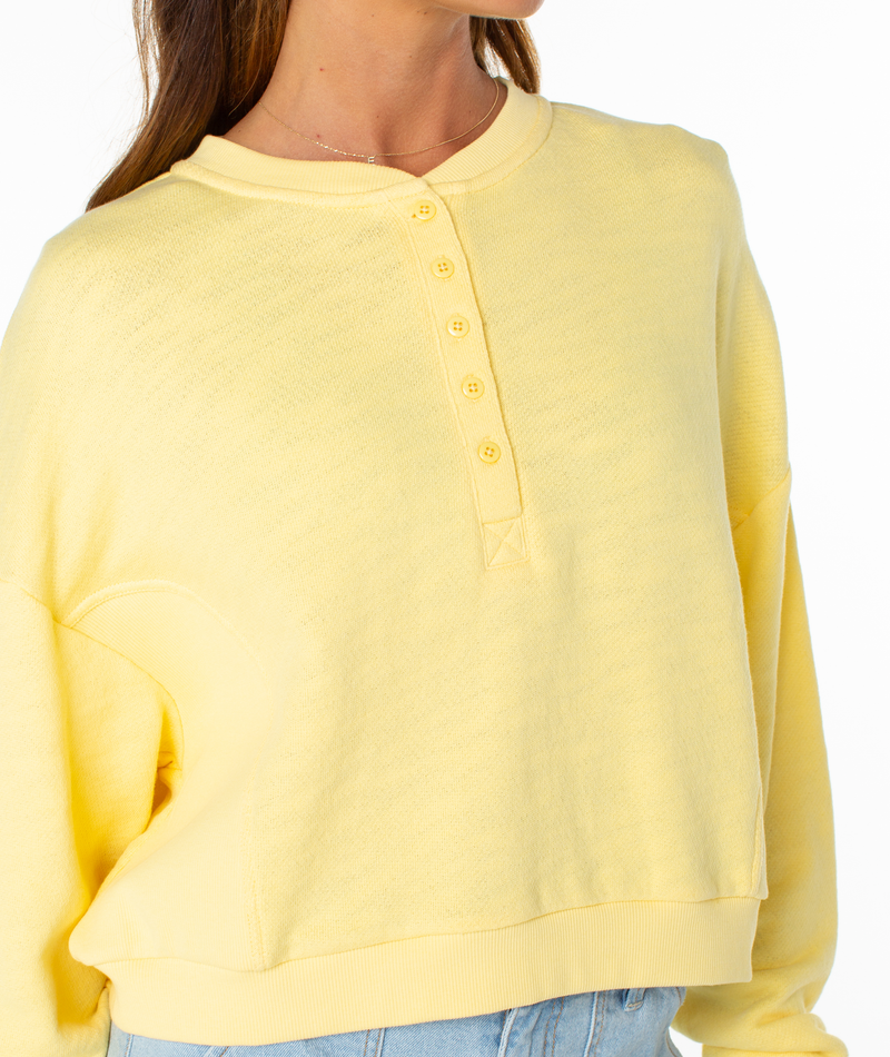 A person wears a light yellow Roxy Overcast Hoodie with a button front, styled with light blue jeans. The cozy cotton crewneck’s relaxed vibe is highlighted as the head is partially out of frame.