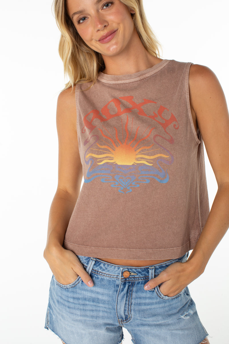 A smiling woman with blonde hair wears a sleeveless Roxy Garrett Wave Tank featuring a colorful sun graphic and blue denim shorts, standing with her hands in her pockets against a white background.