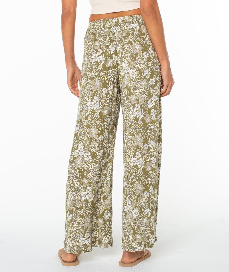 A person is shown from the back wearing Roxy's Midnight Ave Pant in olive green with a white floral pattern, paired with a beige top and sandals against a plain white background.