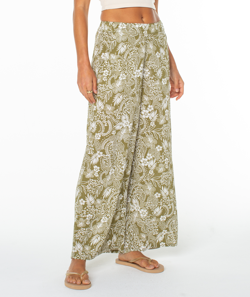 A person wears Roxy's Midnight Ave Pant—olive green, high-waisted with a white floral pattern—paired with a beige crop top and tan sandals, standing against a plain white background.
