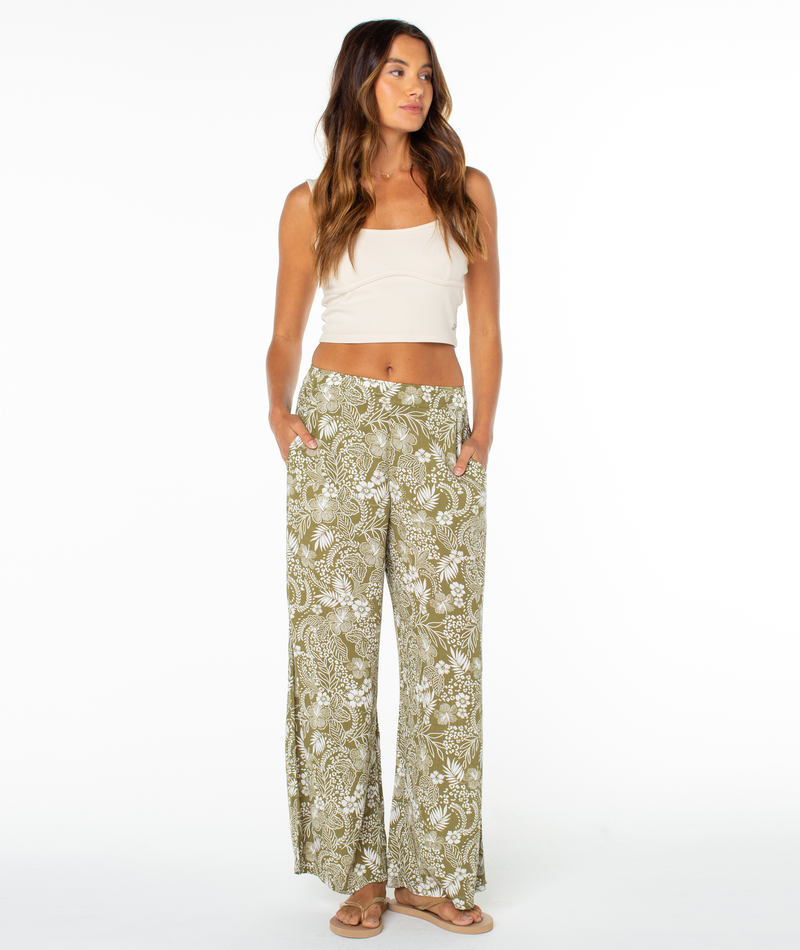 A woman stands against a white background wearing the Roxy Midnight Ave Pant—olive green, high-waisted, floral print lounge pants—with a cream crop top and sandals. She looks slightly to the side with her hands in her pockets.