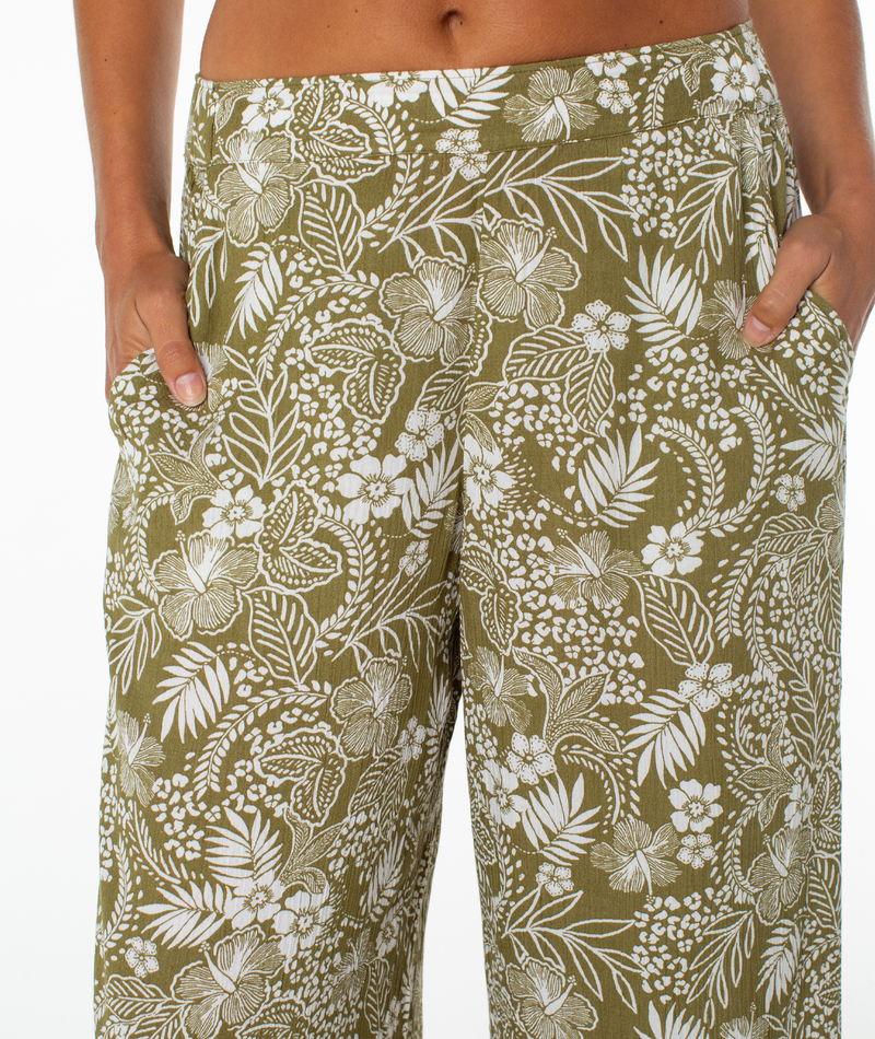 Close-up of a person wearing Roxy’s Midnight Ave Pant—olive green, high-waisted with white floral and leaf patterns—standing with hands in pockets against a white background; perfect for relaxed coastal nights.