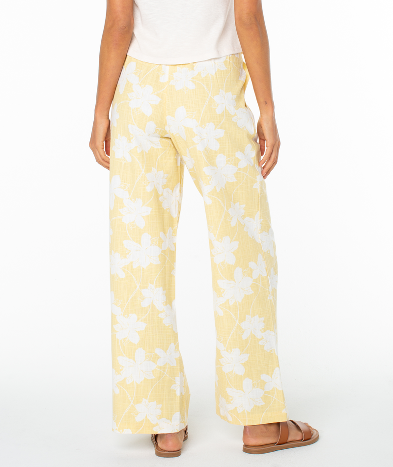 A person faces away against a plain white background, wearing Roxy’s Mavericks Trouser in light yellow with a white floral pattern, paired with a white top and brown sandals.