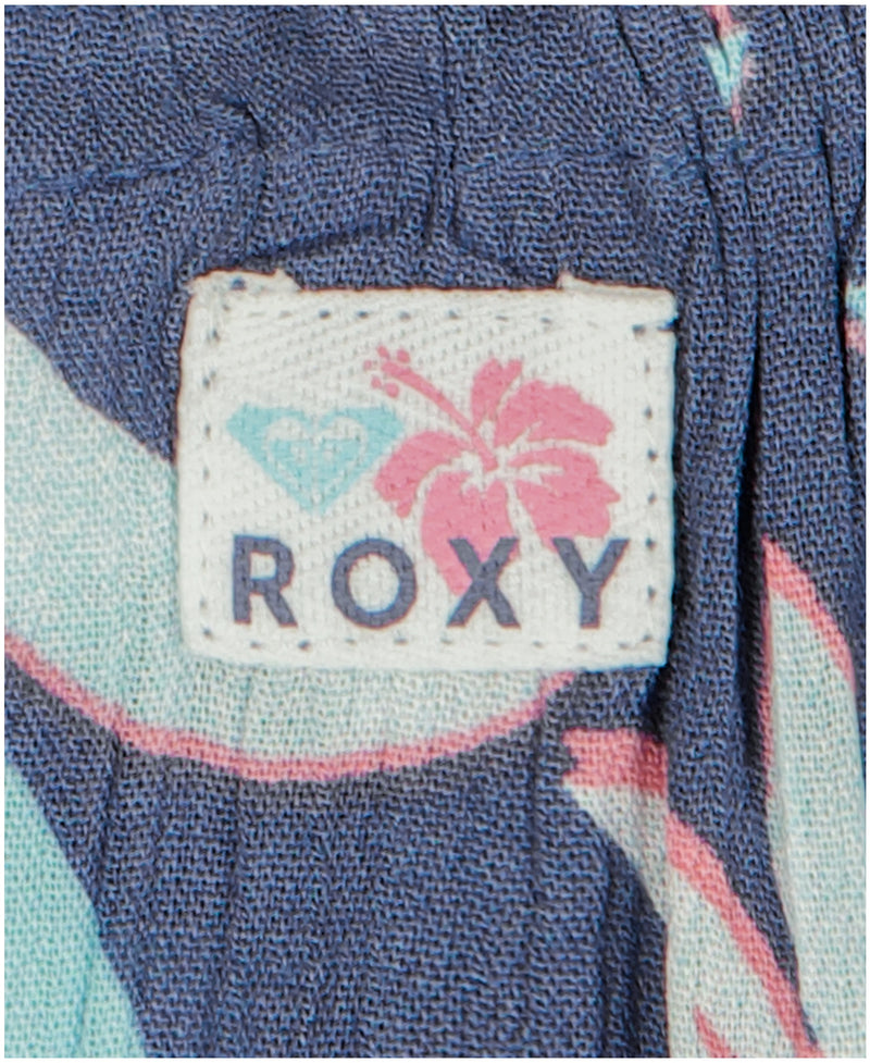 A close-up of a fabric tag on the Girls' Hibiscus Hype Dress by Roxy, featuring the brand name, a pink hibiscus flower, and a blue heart-shaped logo on a patterned blue background.