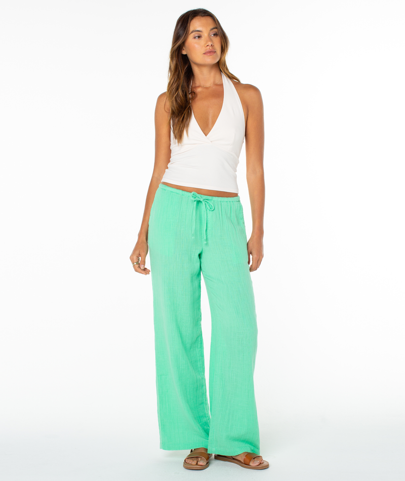 A woman stands against a white background wearing the Roxy South Shore Halter Tank, loose mint green drawstring pants, and brown sandals. She has long hair, a relaxed expression, and her arms at her sides.