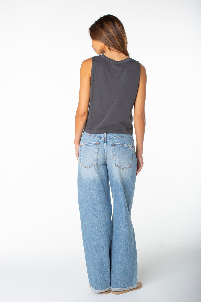 Woman standing with her back to the camera, wearing a sleeveless dark gray Roxy Windy Palm Tank and light blue wide-leg jeans, showing effortless tomboy style against a plain white background.