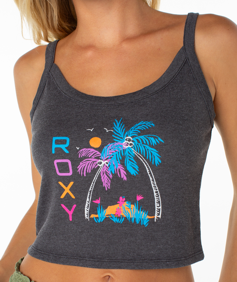 A person wearing the Roxy On The Beach Wanderer Tank—a dark gray cotton blend cropped top with colorful “ROXY” text and a tropical print of palm trees, flowers, a hut, and yellow sun—makes for a true wardrobe staple.