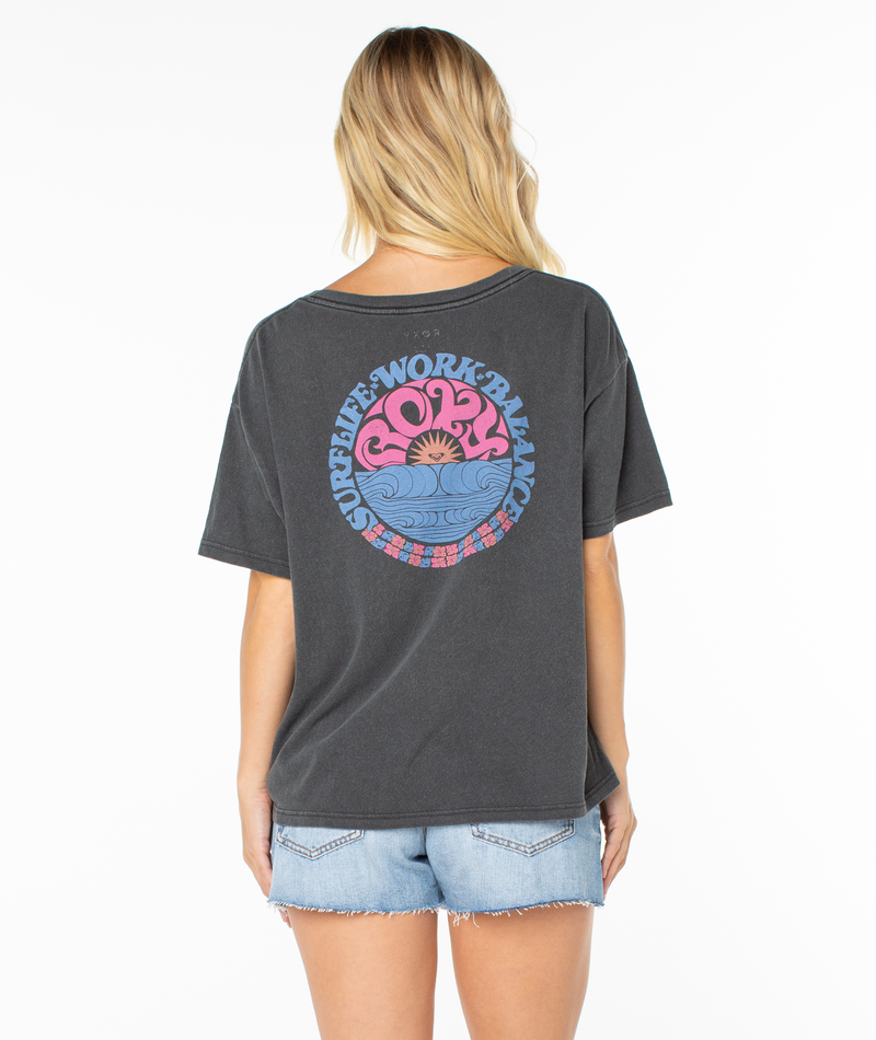 A woman with long blonde hair is seen from the back, lounging in a dark gray Roxy Oversized Vneck Tee with a colorful circular sunset graphic and "SUPREME*WORK*BALANCE" text, paired with denim shorts.