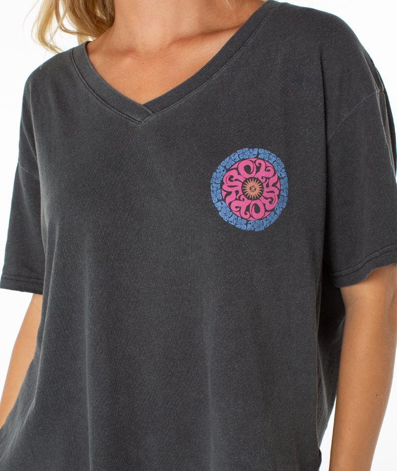 A person lounges in the Roxy Oversized Vneck Tee—dark gray with a vibrant pink and blue circular floral design on the left chest. Only their upper torso is shown against a white background.