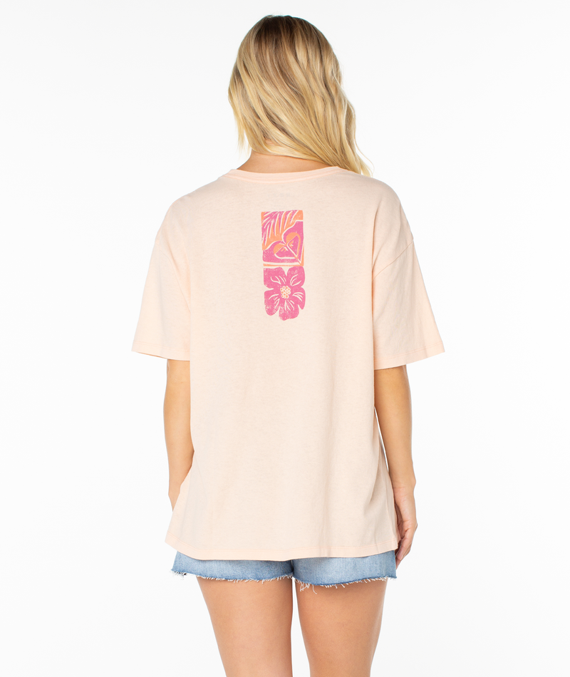 A woman with long blonde hair, facing away from the camera against a white background, wears the Roxy Beach Things Tee—an oversized top with a pink and orange tropical floral graphic on the back—paired with denim shorts.