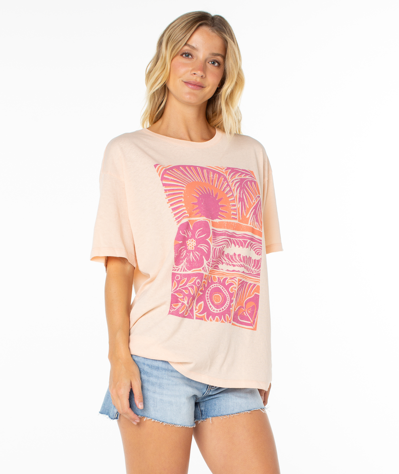 A woman with wavy blonde hair poses casually against a white background, wearing the Roxy Beach Things Tee—an oversized light peach shirt with a pink and orange floral graphic—paired with light denim shorts.