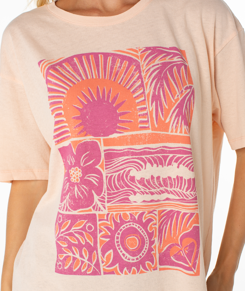 A person wears the Roxy Beach Things Tee, a relaxed drop-shoulder, light peach oversized shirt with pink and orange sun, leaves, flowers, and wave block graphics. Only their upper torso is shown.